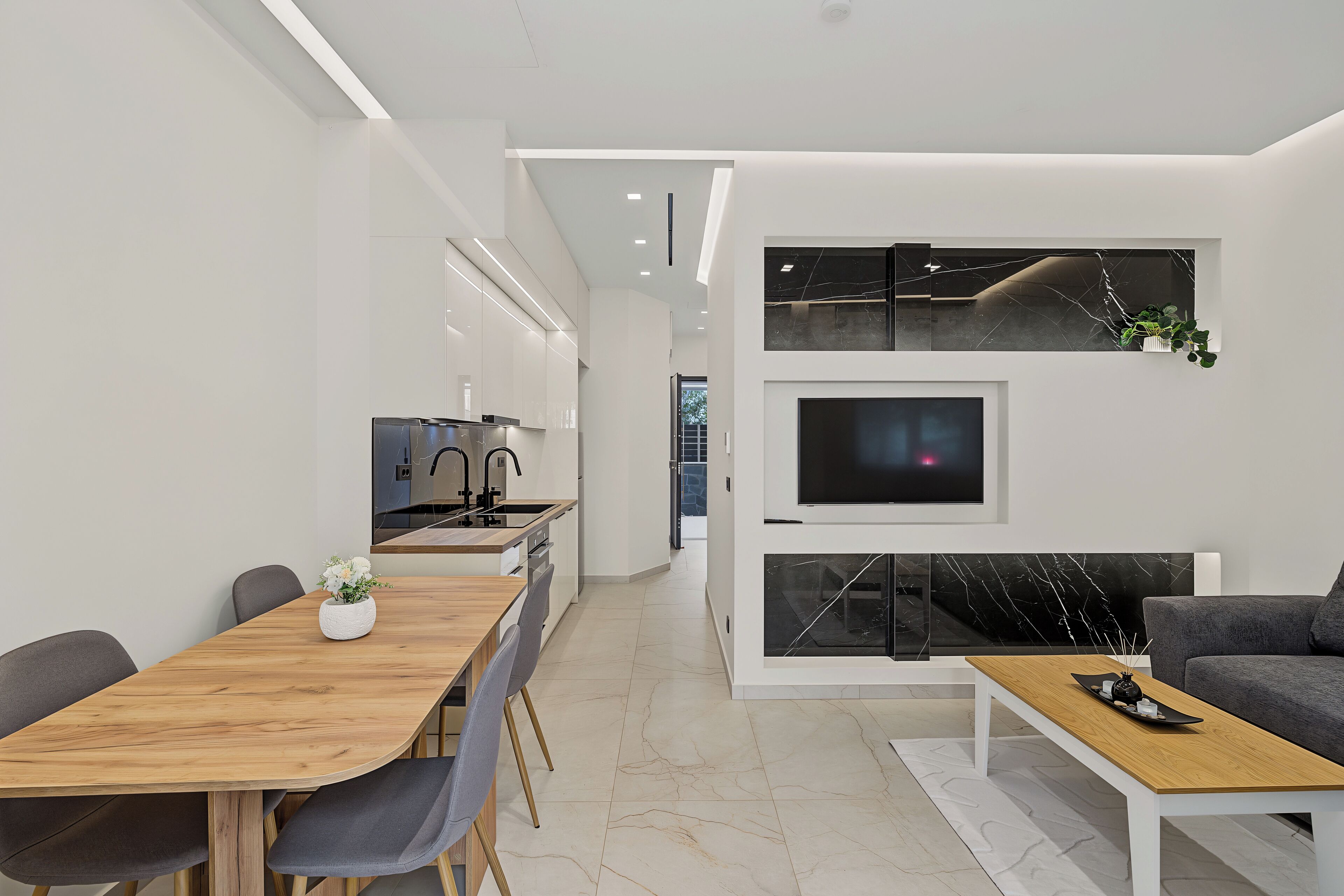 Une cuisine moderne dotée d’un four encastré, un coin repas avec une table et des chaises en bois, et un salon avec un canapé et un téléviseur à écran plat fixé au mur.