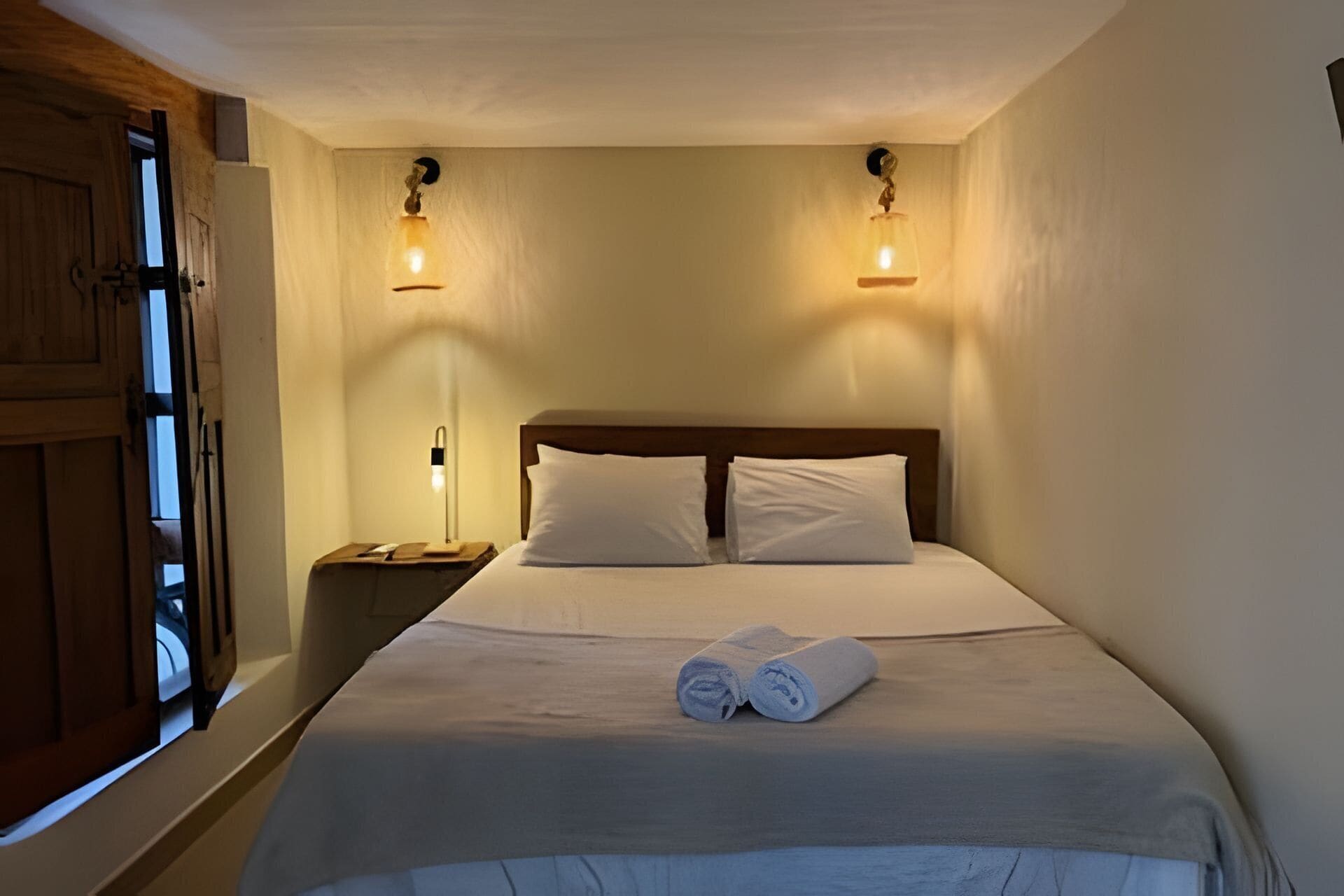 A single bed with white linens and two pillows, a wooden headboard, a nightstand with a lamp, and a window with a view.
