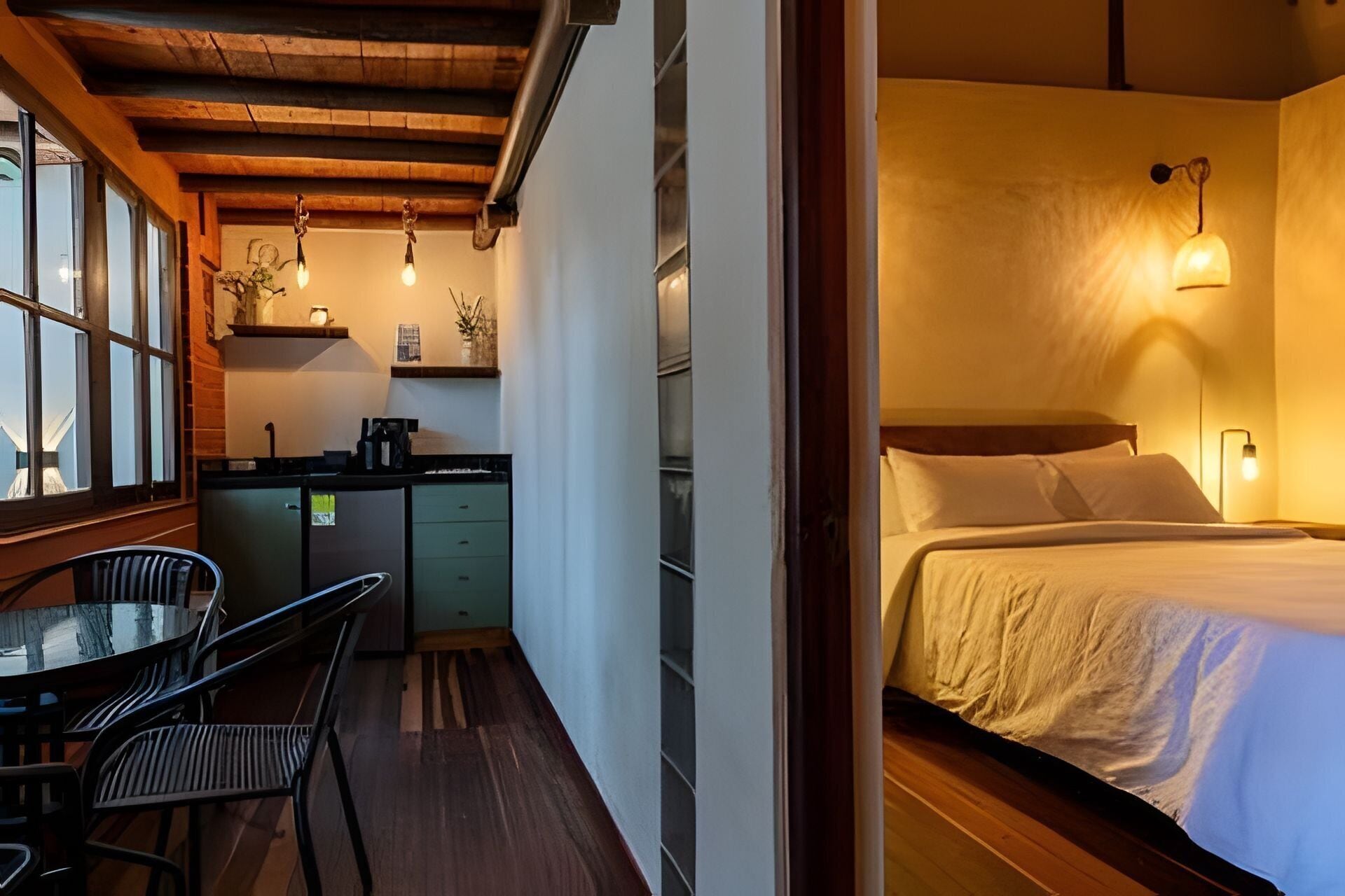 A compact hotel room with a bed, bedside table, and a small kitchenette area.