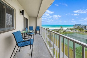 Ein Balkon mit Tisch und Stühlen, mit Blick auf einen Strand und einen Tennisplatz.