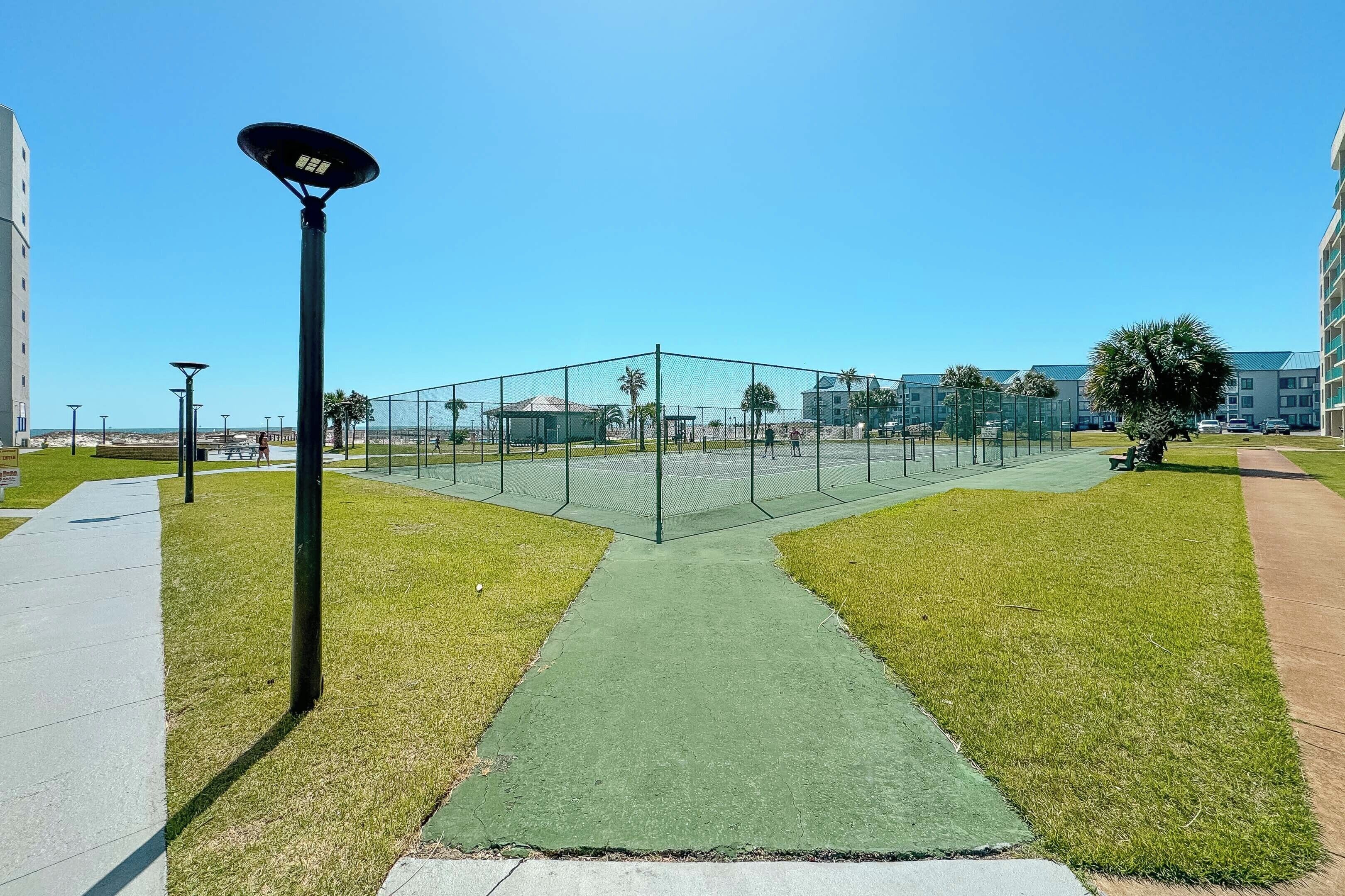 A fenced sports court with a green surface, surrounded by grass and a sidewalk.