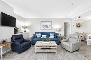 A modern living room with a blue sofa, two armchairs, a coffee table, and a flat-screen TV mounted on the wall. - Beachfront 2BR, Stunning Gulf Views, Indoor/Outdoor Pools, Hot Tub, Tennis Court (Gulf Shores)