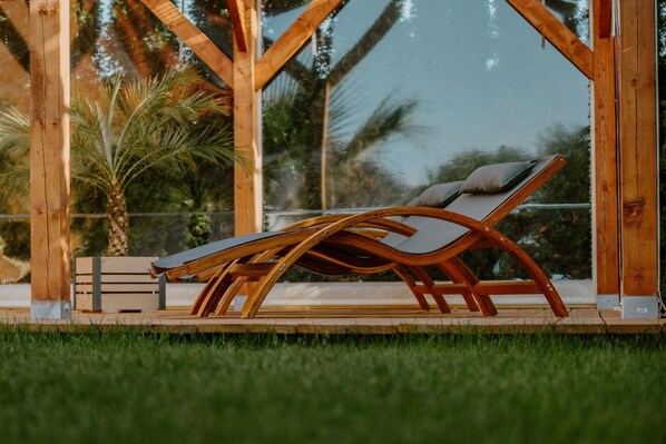 A wooden lounge chair with a cushion, positioned under a glass structure with a view of palm trees and a body of water.