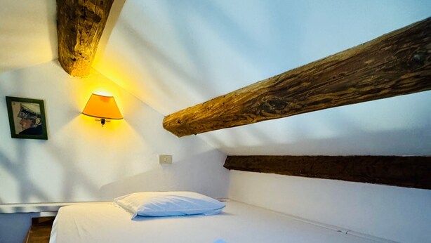 A single bed with white linens and a folded towel, a wall-mounted lamp, and a wooden beam above the bed. - Rialto Vacations with Air Conditioning and Shared Kitchen (Venice)