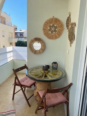 Ein Balkon mit einem runden Glastisch, zwei Holzstühlen mit rosa Kissen und dekorativen Wandbehängen.