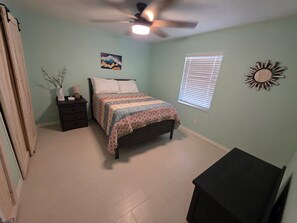Um quarto com cama, mesa de cabeceira, ventilador de teto, janela com persianas e um relógio de parede.