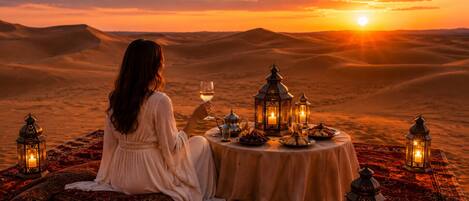 Una persona sentada en un espacio con alfombra, con una mesa dispuesta para una comida, rodeada de faroles, en un paisaje desértico al atardecer.