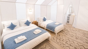 Una habitación con tiendas de campaña, con dos camas, cada una con una manta azul y almohadas blancas, una mesita de noche y una lámpara colgante.