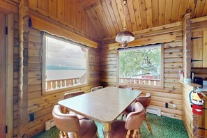 A wooden cabin interior with a dining table and chairs, large windows, and a ceiling fan.