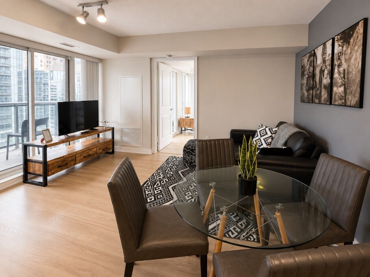 A modern living room with a glass dining table, leather sofas, a flat-screen TV, and a large window offering a city view.