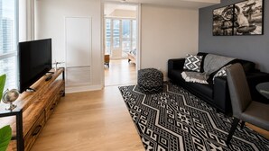 A modern living room with a flat-screen TV, a black leather sofa, a patterned rug, and a city view through large windows.