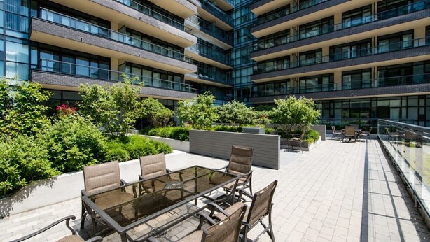 A modern outdoor seating area with a glass table and chairs, surrounded by lush greenery and a multi-story building with balconies. - Bright 1BR at Motion | Prime Downtown Location (Toronto)