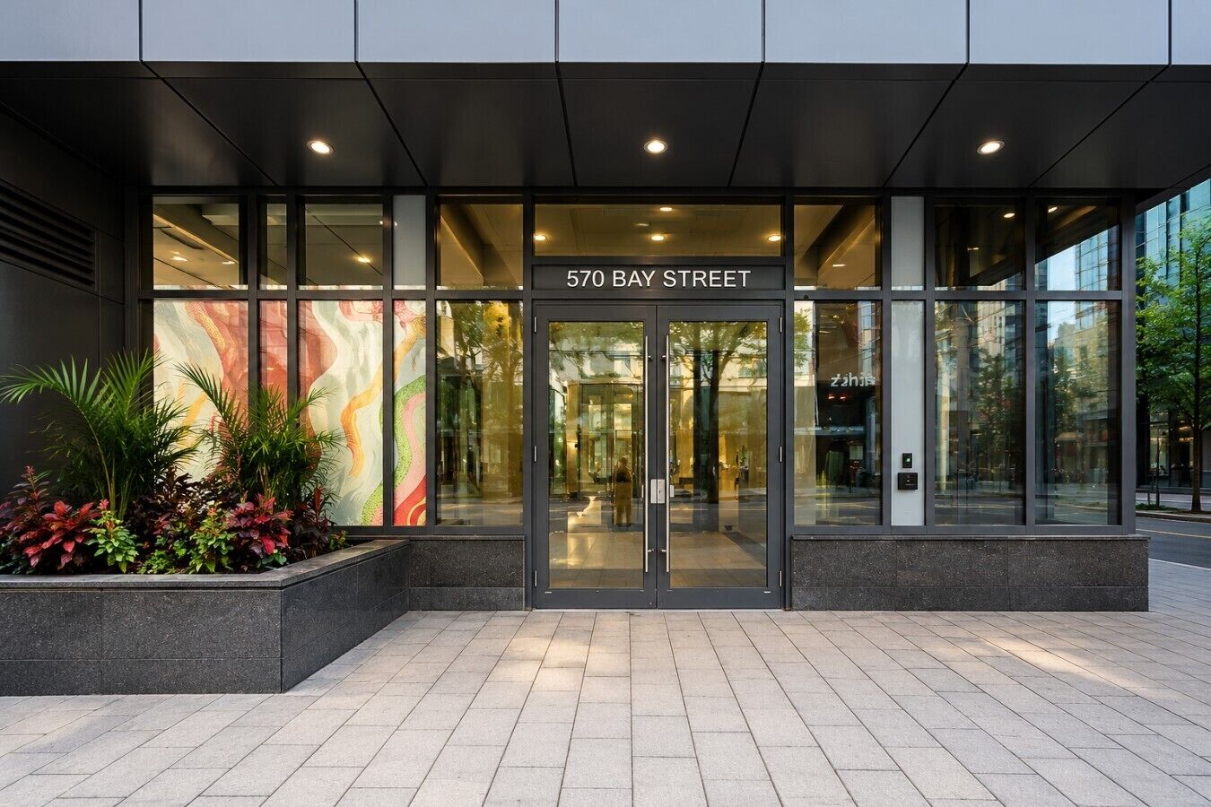 A modern building entrance with a glass facade, a sign reading '570 BAY STREET,' and a landscaped area with plants.