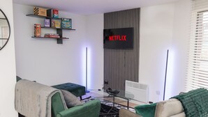 Ein modernes Wohnzimmer mit einem Flachbildfernseher, der 'NETFLIX' zeigt, einem Bücherregal mit Brettspielen und einem Sofa mit Decke.