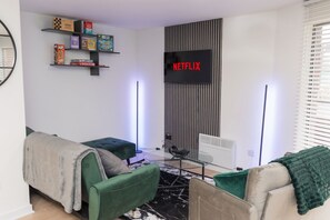 A modern living room with a flat-screen TV displaying 'NETFLIX,' a bookshelf with board games, and a sofa with a blanket. - Game Night Apartment | Walk to City Centre (Wolverhampton)