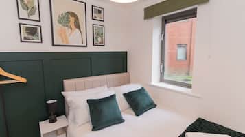 A neatly arranged bedroom with a bed, pillows, a bedside table, a lamp, and framed botanical artwork on the wall.