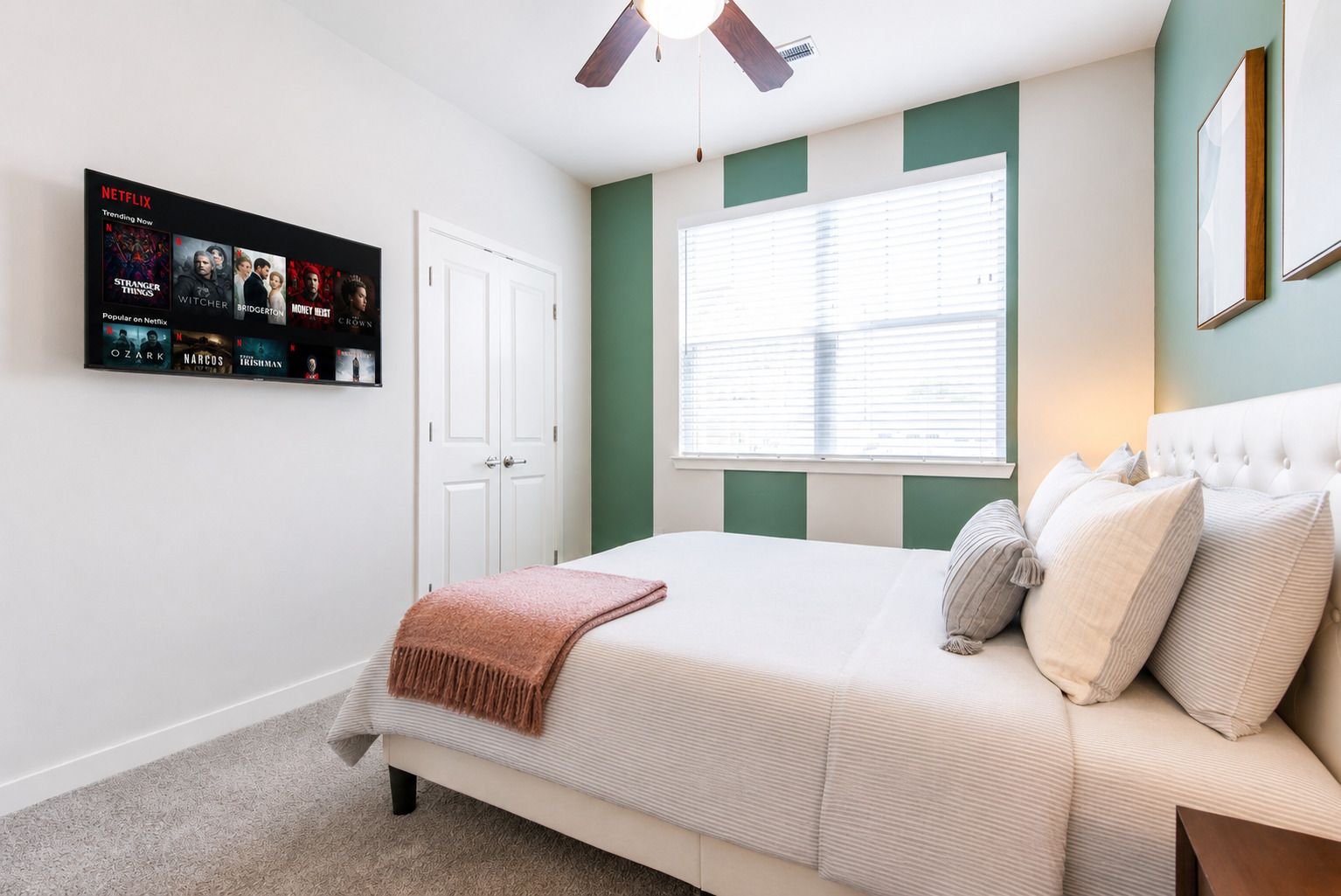 A bedroom with a bed, a TV mounted on the wall displaying Netflix content, a window with blinds, and a ceiling fan.