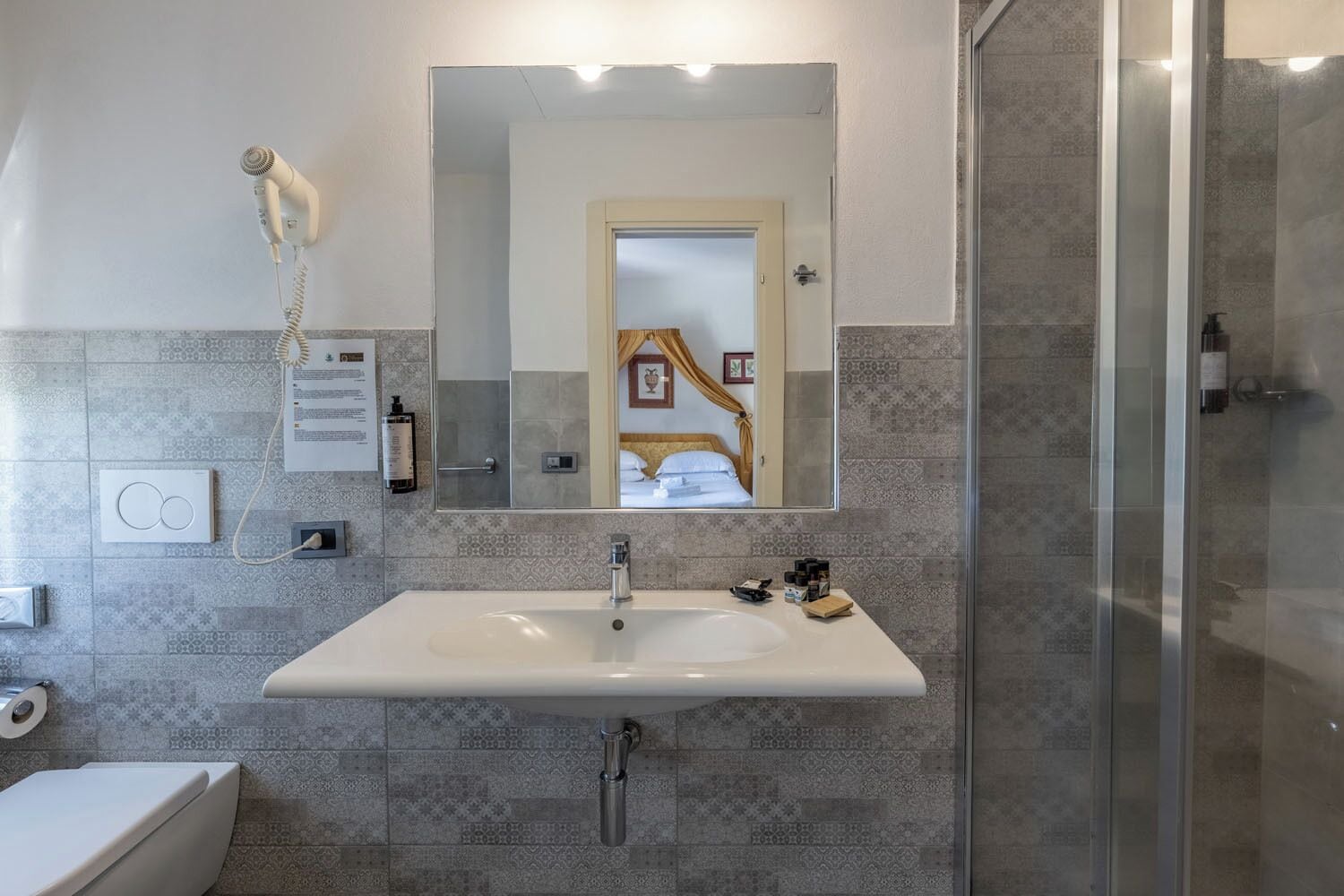 Un baño con un espejo grande, un lavamanos blanco, un box de ducha y un secador de pelo montado en la pared.