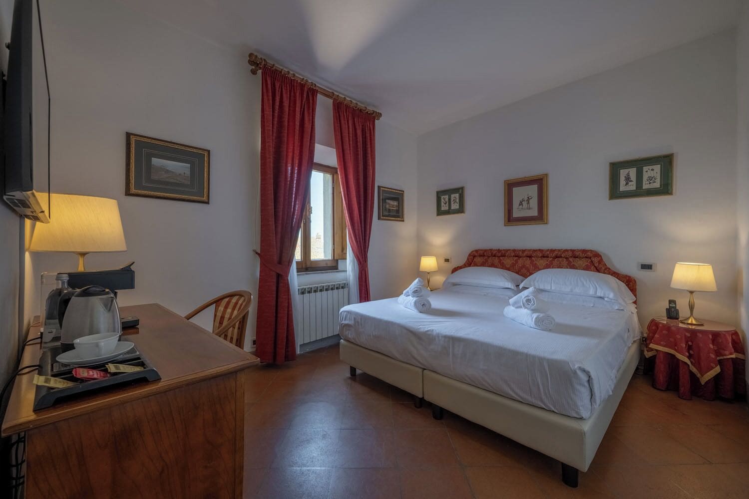 Ein Hotelzimmer mit Bett, Nachttischlampen, einem Schreibtisch mit Kaffeemaschine und einem fenster mit Vorhängen.