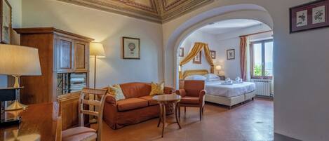 Habitación de hotel con cama, sofá, mesa y una silla. El techo tiene diseños decorativos y una ventana con cortinas.
