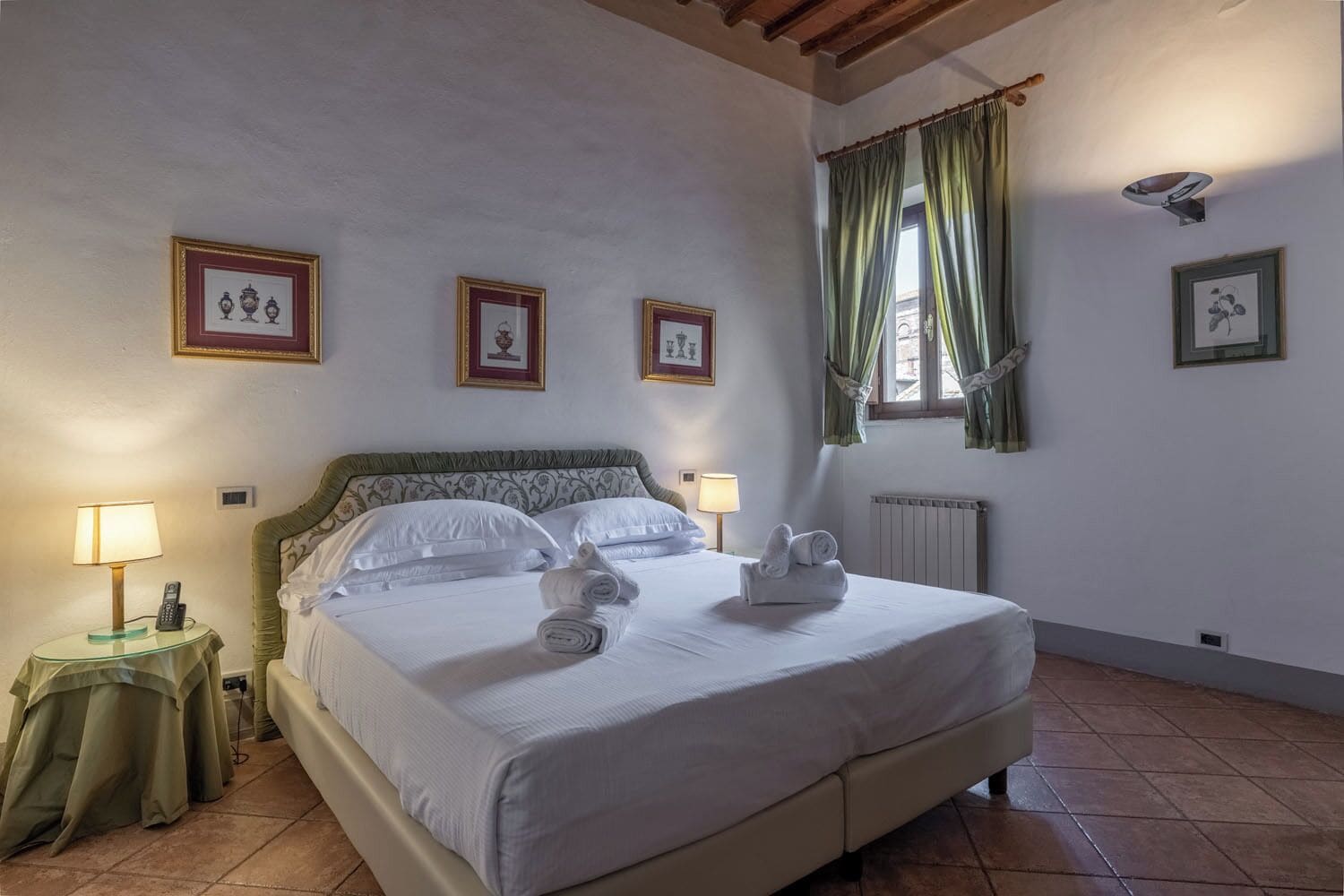 Una cama bien hecha con almohadas y toallas enrolladas, una mesita de noche con lámpara, y cuadros en la pared.