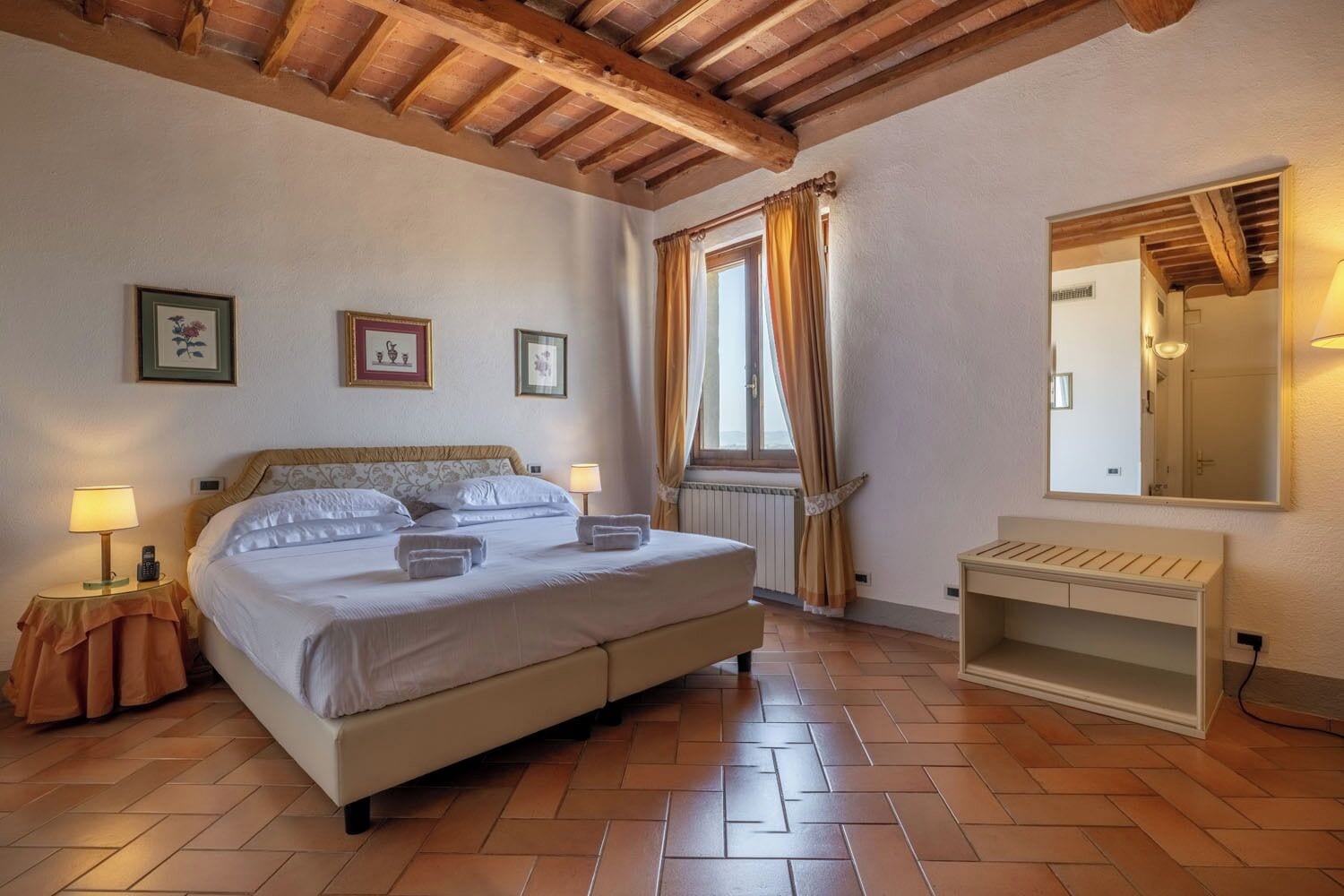 A bedroom with a bed, bedside lamps, a bedside table, a mirror, and a window with curtains.