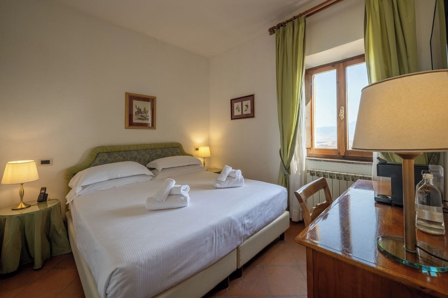 A hotel room with a bed, two bedside lamps, a table with a lamp, and a window with curtains.