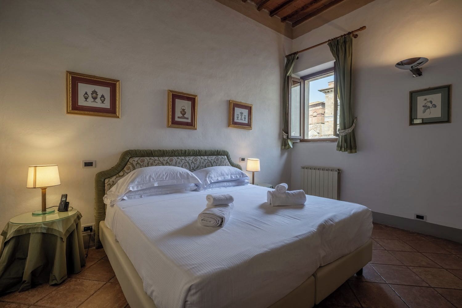 Una camera d'albergo con un letto, due abat-jour, un telefono, un termosifone e quadri appesi alle pareti.