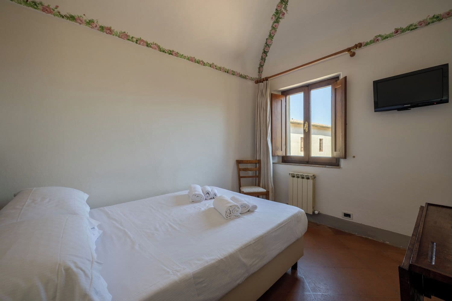 Quarto de hotel com cama, cadeira, janela com cortinas, radiador e televisão fixada na parede.