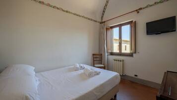 Quarto de hotel com cama, cadeira, janela com cortinas, radiador e televisão fixada na parede.