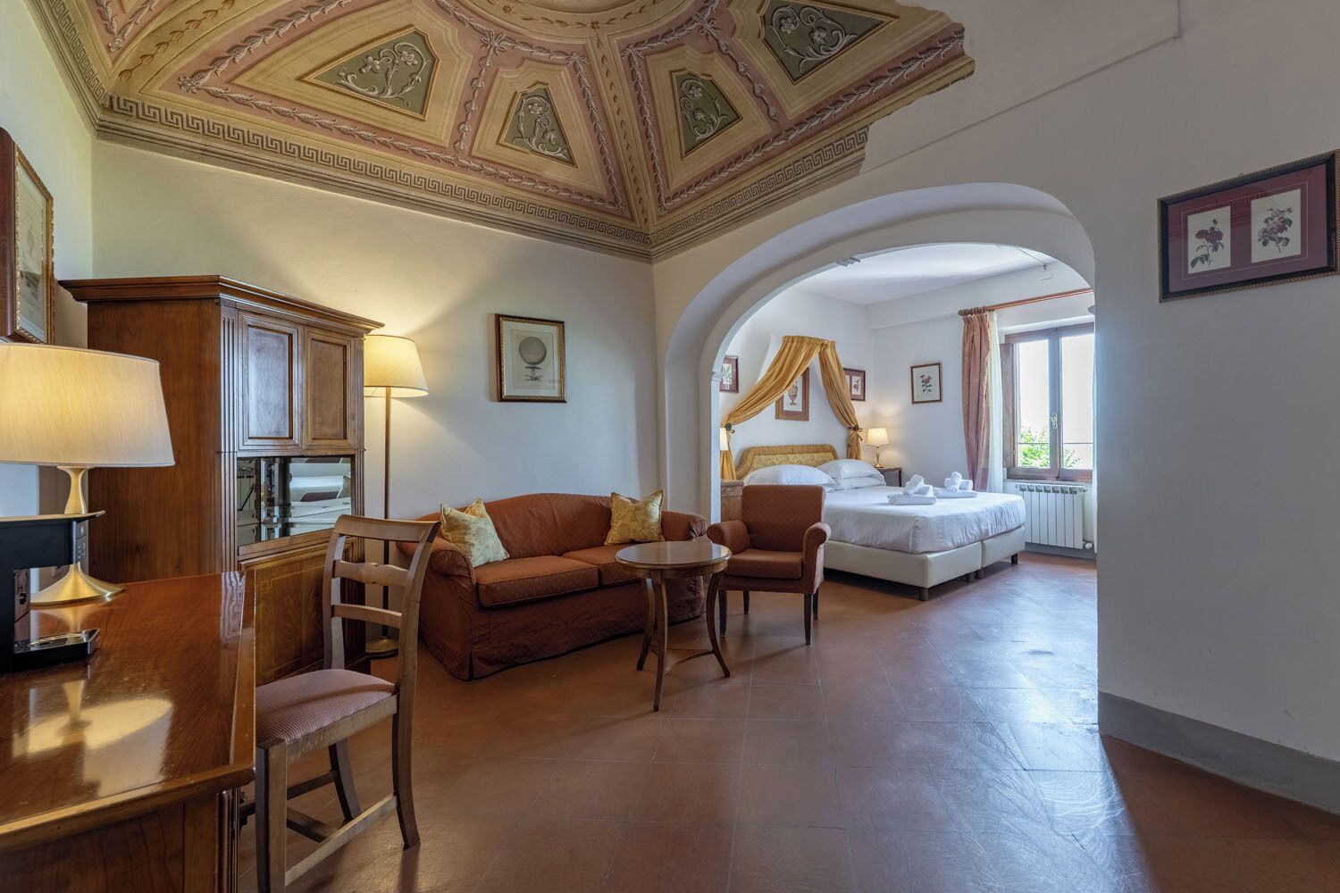 Habitación de hotel con cama, sofá, sillón, escritorio y lámpara. El techo presenta diseños decorativos, una ventana con cortinas y cuadros en la pared.