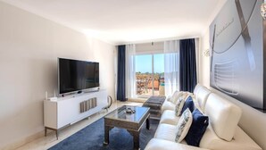 Ett modernt vardagsrum med en platt-TV, en vit soffa, ett soffbord och ett stort fönster med gardiner.