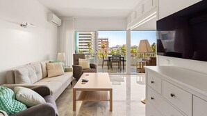 A modern living room with a sofa, a coffee table, and a TV. There is a balcony with a table and chairs, and a city view. - RDR568 - Charming 1 Bed Apt in Jardines del Mar (Marbella)