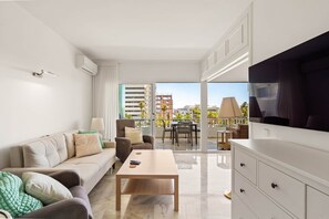 Ein modernes Wohnzimmer mit einer Couch, einem Couchtisch und einem Fernseher. Ein Balkon mit Tisch und Stühlen bietet Blick auf die Stadt.