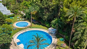 A large circular pool surrounded by palm trees and lounge chairs, with a smaller pool and a building in the background. - RDR568 - Charming 1 Bed Apt in Jardines del Mar (Marbella)