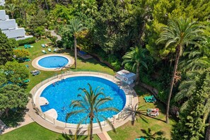 Ein großer, runder Pool, umgeben von Palmen und Liegestühlen, mit einem kleineren Pool und einem Gebäude im Hintergrund.