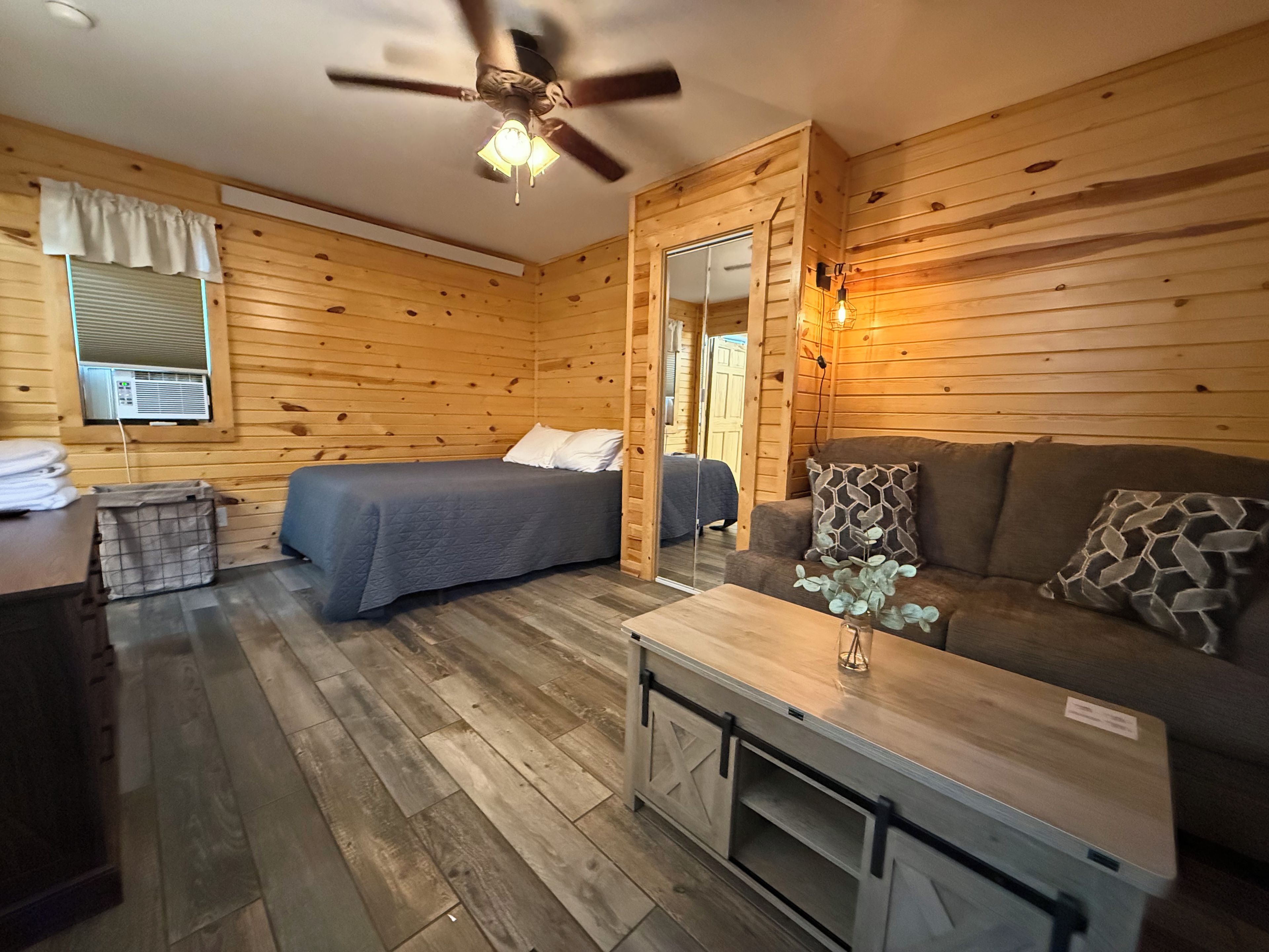 A room with a bed, a sofa, a small table, a ceiling fan, and a window with blinds.