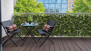 Ein Balkon mit zwei Stühlen, einem kleinen Tisch, einer Topfpflanze und Blick auf ein Gebäude mit vielen Fenstern.