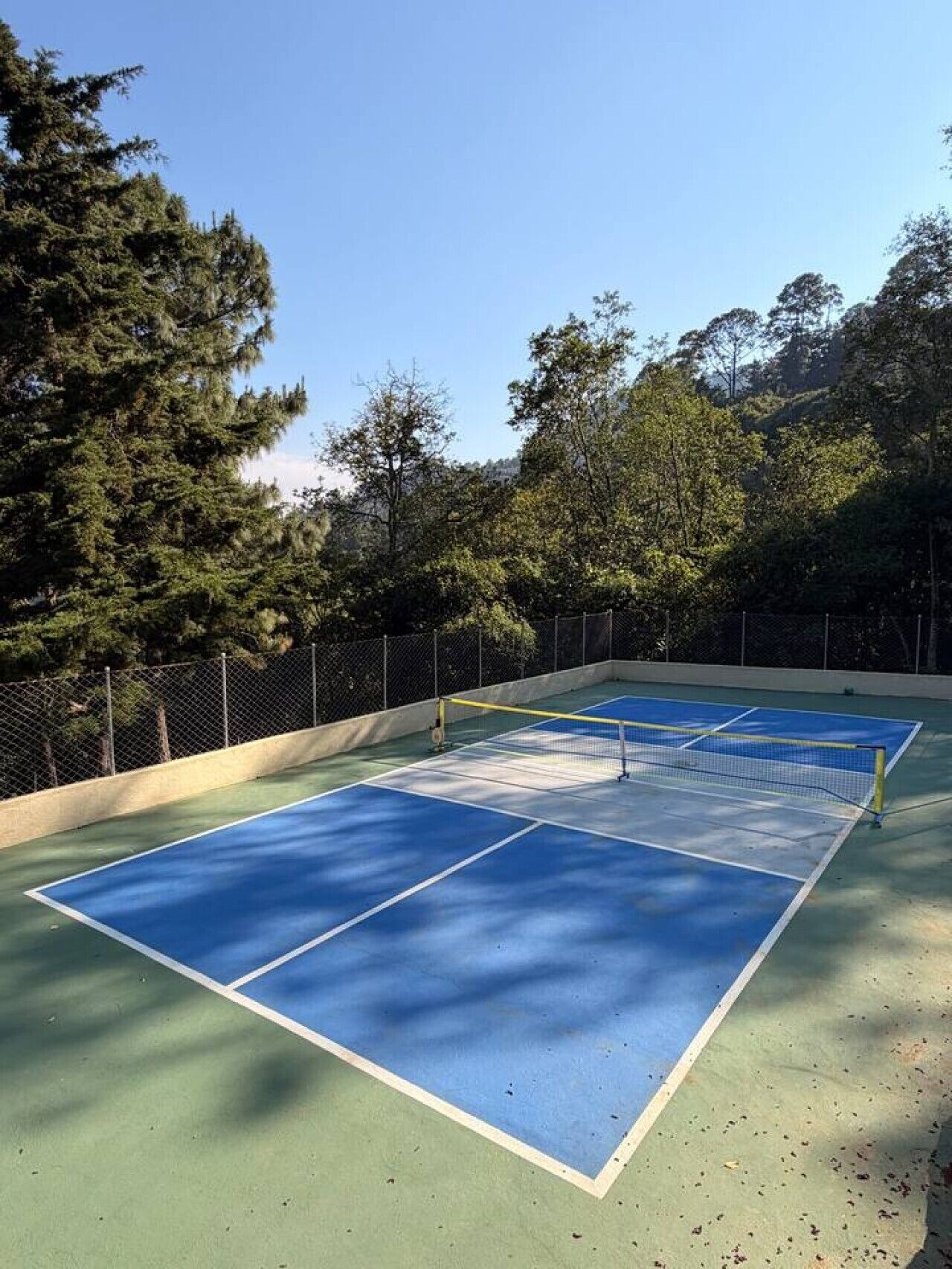 Una cancha de tenis al aire libre con una red, rodeada por una cerca de malla metálica y árboles.