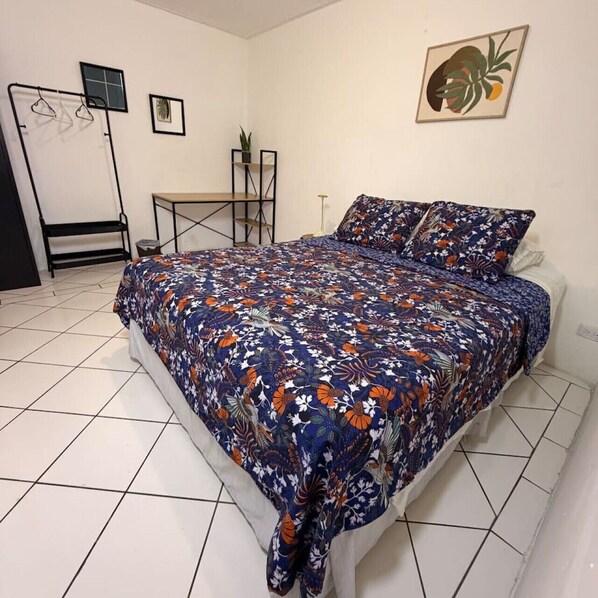 A bedroom with a bed, a desk, a chair, a potted plant, and a framed picture on the wall.