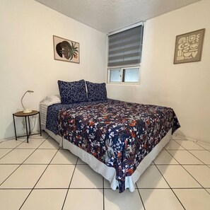 A bedroom with a bed, a bedside table, a window with blinds, and two wall decorations.