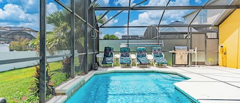 Une piscine dotée d’une structure en verre, de chaises longues et d’un espace barbecue.