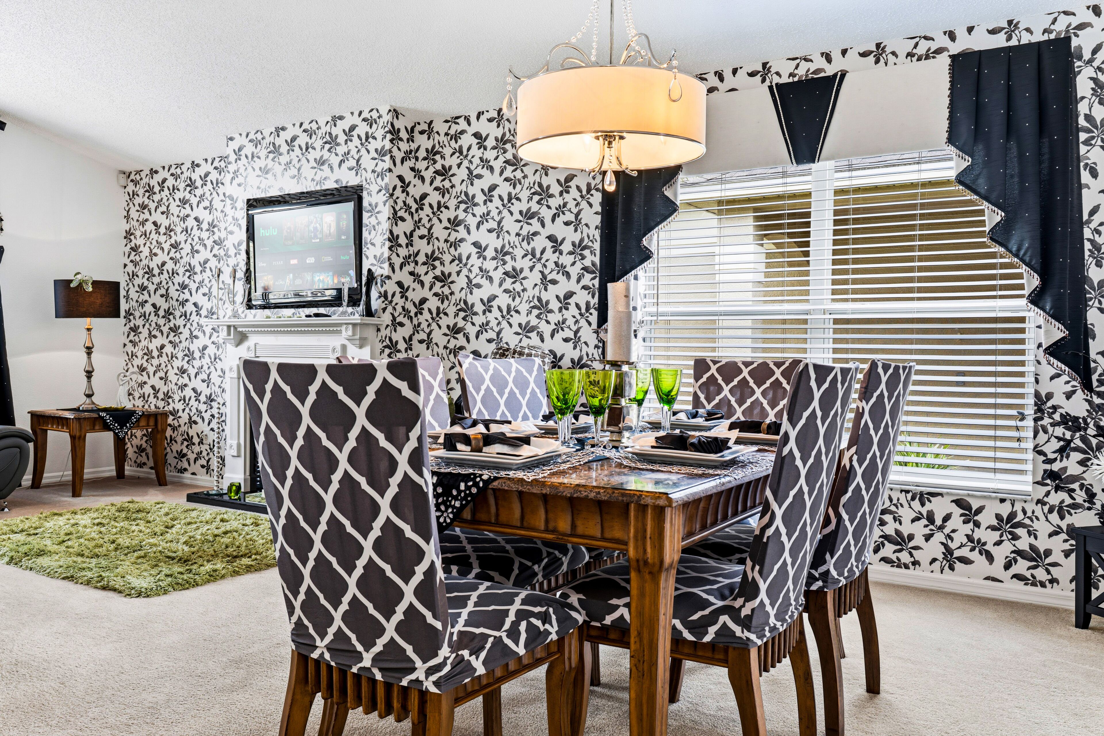 Un coin repas avec une table dressée pour quatre personnes, entourée de chaises à motifs, un téléviseur fixé au mur et une fenêtre avec des stores.