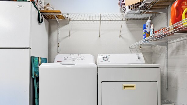 A compact laundry area with a front-loading washer and dryer, a shelf with cleaning supplies, and a small trash bin. - 4 BDR South-Facing Disney Retreat (Kissimmee)