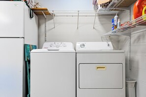 Un área de lavandería compacta con lavadora y secadora de carga frontal, un estante con productos de limpieza y un pequeño cubo de basura.