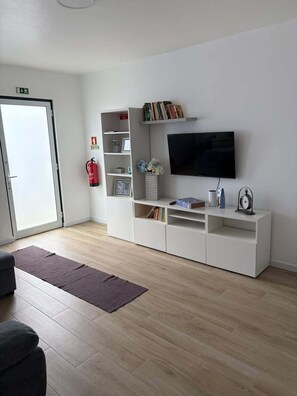 Ein modernes Wohnzimmer mit einem an der Wand befestigten Flachbildfernseher, einer weißen Unterhaltungsanlage mit Büchern und Dekorationsobjekten, einem Feuerlöscher an der Wand und einer Glastür, die nach draußen führt.