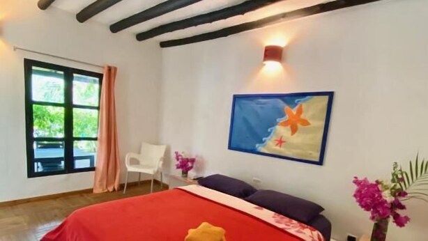 A bedroom with a large bed, a nightstand, a chair, a window with curtains, and a painting on the wall. - Italcaribe Village (Paraguachí)