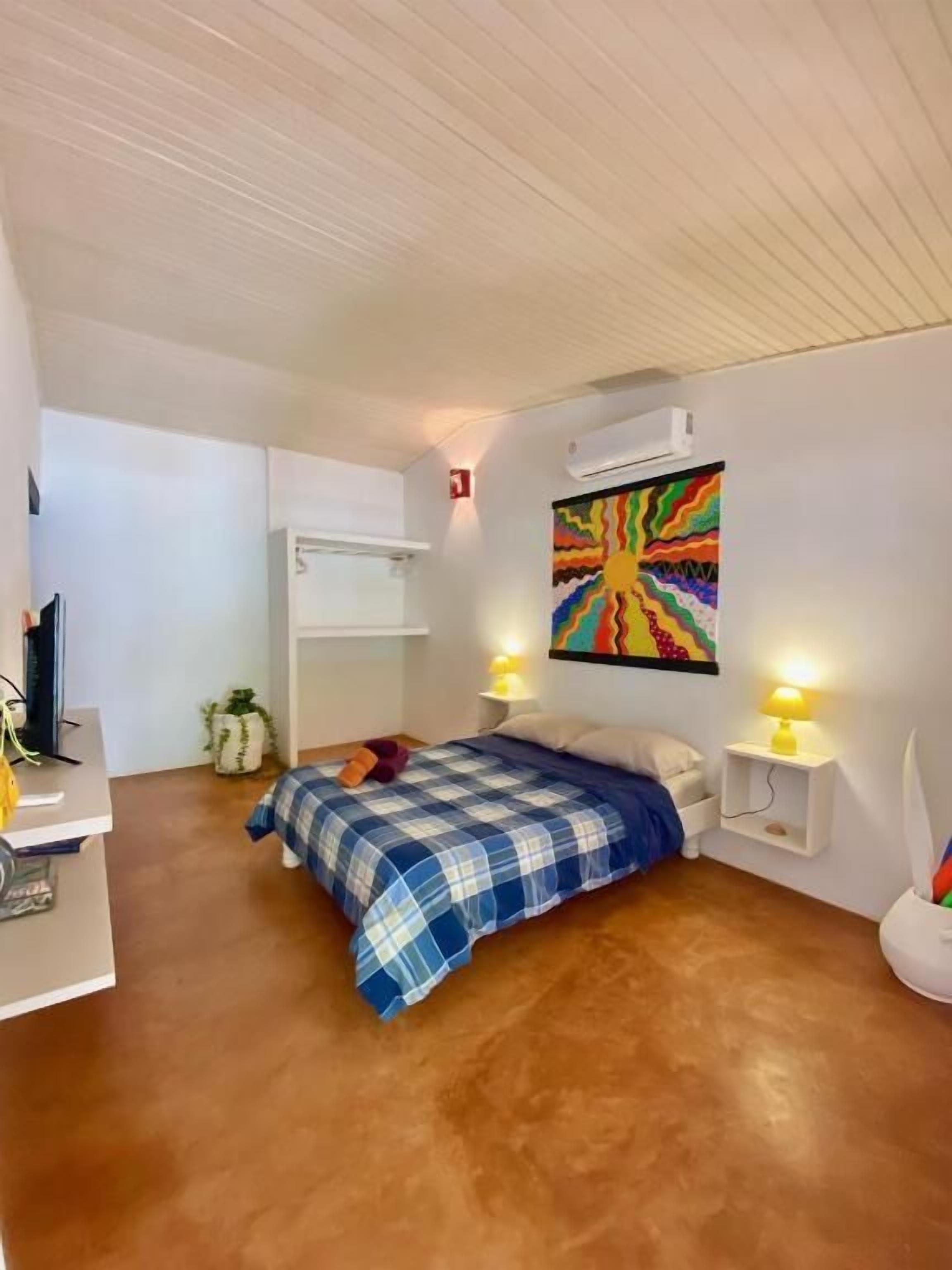 Quarto com cama, luminárias de cabeceira, ar condicionado fixado na parede e uma pintura abstrata colorida.