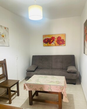 A living room with a grey sofa, a wooden table with a map tablecloth, a wooden chair, and a painting of red flowers on the wall. - Margarita Penthouse Conil Center – Balcony & Air Conditioning (Conil de la Frontera)