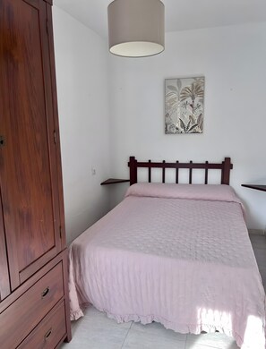 A bedroom with a wooden wardrobe, a bed with a pink bedspread, a hanging light fixture, and a wall-mounted artwork. - Margarita Penthouse Conil Center – Balcony & Air Conditioning (Conil de la Frontera)